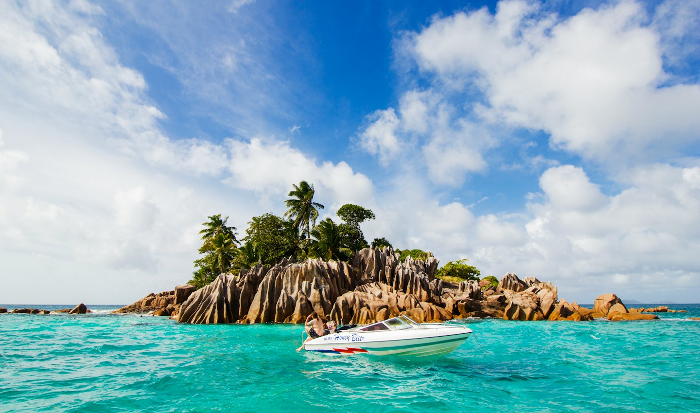 A speedboat carries two passengers past a tropical island.